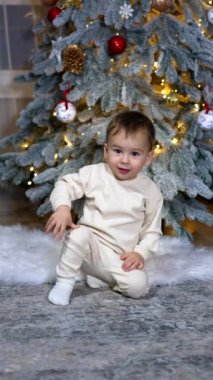 Odada Noel ağacının yanında oturan küçük sevimli melek bebek. Sevimli çocuk sanki bir şey bekliyormuş gibi hareketsiz oturur ve kamera yaklaşırken sevimli bir şekilde gülümser. Dikey video