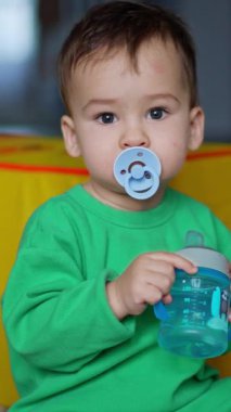 Ağzında emzik olan tatlı, uykucu bir çocuk yerde sakince oturuyor. Bebek elinde suyla şişeyi tutuyor. Dikey video