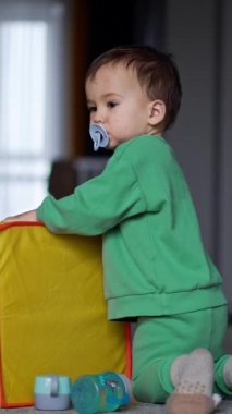 Tatlı çocuk büyük sarı oyuncak kutusunun önünde diz çöküyor. Emziği olan çocuk ayağa kalkıp yürür. Dikey video