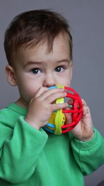 Koyu renk saçlı çocuk ağzına oyuncak dayıyor. Küçük bebek odada oyuncaklarla oynuyor. Dikey video