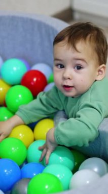 Tatlı çocuk, içi renkli oyuncaklarla dolu kuru havuzda oynuyor. Küçük çocuk içeride oynuyor. Üst Manzara. Dikey video