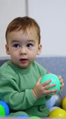 Beyaz tatlı çocuk kuru havuzun içinde oturuyor. Bebek bir top seçiyor, tutuyor ve kameraya bakıyor. Dikey video