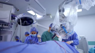 Gelişmiş ekipmanlı modern cerrahi odası. Cerrahlar mikroskoba bakan bir hastayı ameliyat ederler. Dişi hemşire hazırlık araçlarına yardım eder.