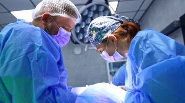 Kadın cerrah ameliyatta hastanın üzerine eğiliyor. Doktor elektrikli alet için aleti değiştirir. Erkek asistan ameliyata yardım ediyor. Düşük açı görünümü.