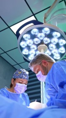 Hastanın üstündeki yuvarlak lambanın altında iki tıp arkadaşı duruyor. Doktorlar tiroid üzerinde metal aletler kullanırlar. Düşük açı. Dikey video.