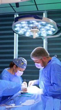 Sıhhiyeciler tiroit ameliyatı yaparlar. Baş kadın cerrah ve yardımcı doktor parlak yuvarlak lamba altında çalışıyorlar. Dikey video.