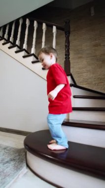Çıplak ayaklı beyaz çocuk, odanın merdivenlerinde dikiliyor. Neşeli erkek bebek enerjik bir şekilde zıplar ve sonra odadan kaçar. Düşük açı. Dikey video.