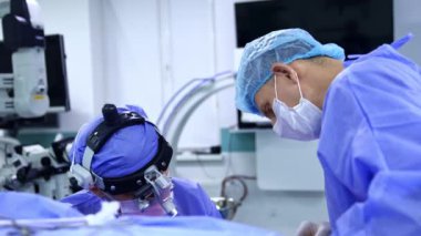 Hastanın başında oturan erkek cerrah cerrahi hoparlör takıyor. Beyaz maskeli doktor hastaya doğru eğildi..