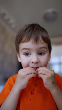 Çilek yiyen sevimli bir erkek bebek. Meyve suyu çocuğun çenesinden akıyor ve tişörtüne damlıyor. Kapatın. Küçük çocuk kirli ellere bakar ve onları kameraya gösterir. Dikey video.