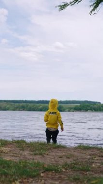 Sarı kazak ve siyah pantolon giyen çocuk gölde duruyor. Küçük çocuk çakıl taşlarını suya atıyor. Dikey video.