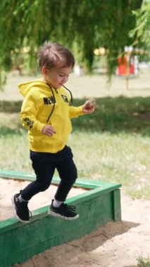 Tatlı çocuk kum havuzundan çıkıyor. Küçük çocuk elinde bir taşla göle doğru yürür ve onu suya atar. Dikey video.