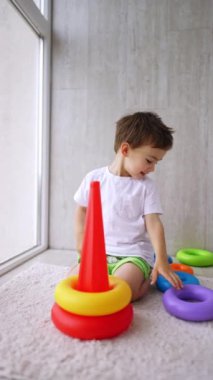 Güzel gülümseyen çocuk piramidin önünde yerde oturuyor. Toddler evde bir oyuncakla oynuyor. Dikey video.