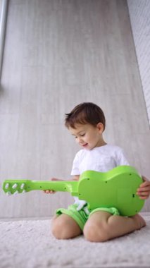 Beyaz, şirin bir çocuk elinde gitarla yerde oturuyor. Evde oynayan çocuğa düşük açılı bakış açısı. Dikey video.