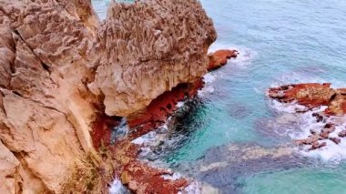 Beyaz köpüklü dalgalar kırmızı kaya oluşumları tarafından sıçrıyor. Deniz kıyısındaki dikenli dağların zirvesi..