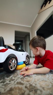 Kafkasyalı küçük çocuk yerde, büyük oyuncak arabasının yanında yatıyor. Meraklı çocuk onların dönüşünü izlemek için otomobilin çarklarını döndürür. Dikey video.