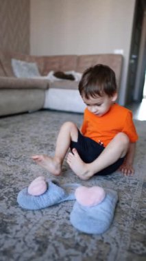 Turuncu tişörtlü güzel erkek bebek yerde oturuyor ve büyük annenin terliklerini çıkarıyor. Çocuk yerde sürünüyor ve bir oyuncağı fark ediyor. Dikey video.