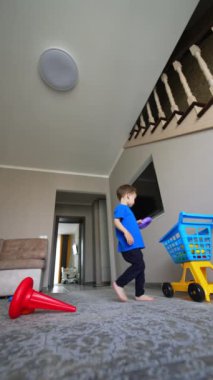 Sevimli erkek bebek oyuncaklarını toplayarak odanın yanından geçiyor. Çıplak ayaklı çocuk arabaya oyuncak topluyor. Düşük açı. Dikey video.