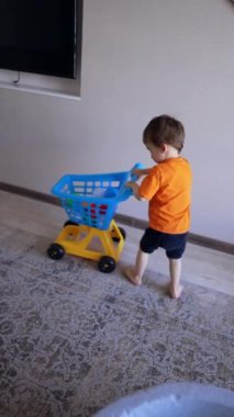 Turuncu tişört giyen ve siyah şortlu tatlı çocuk oyuncak arabasını itiyor. Çocuk neşeyle koridorda koşup başka bir odaya giriyor. Dikey video.