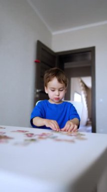 Mavi tişörtlü sevimli beyaz çocuk masasında bulmacalarla oynuyor. Neşeli çocuk parçaları birleştiriyor, ellerini masaya vuruyor ve odadan kaçıyor. Dikey video.