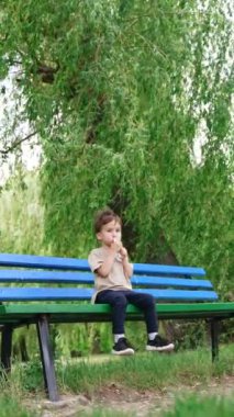 Ağacın altındaki bankta oturan erkek çocuk elinde oyuncak bir çantayla. Bebek yürürken bir şeyler atıştırıyor. Düşük kapsama alanı. Dikey video.