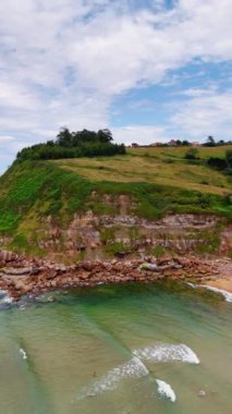 İnsanlar kumsaldaki şeffaf sularda duruyor. Ayağının altında kaya yığını olan kaya oluşumu ve üzerinde bazı evler. İspanya, Avrupa. Dikey video.