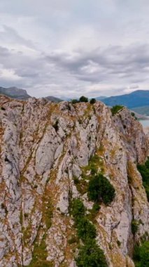 Gri bir kayaya yakın uçuyor ve bitkiler yetişiyor. Arka planda dağların arasında saklanan muhteşem mavi göl manzarası. Gökyüzünde dramatik bulut manzarası. Dikey video.