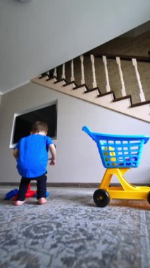 Mavi tişörtlü beyaz bebek oyuncakları arabaya topluyor. Evde tek başına oynayan çocuğa düşük açılı bakış açısı. Dikey video.
