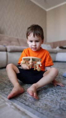 Sevimli erkek bebek elinde tahta bir oyuncakla yerde oturuyor. Çocuk bir oyuncağın eline verip ona hafifçe vurmasına şaşırmış gibi görünüyor. Dikey video.