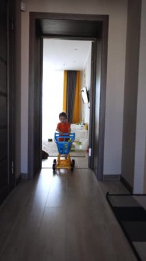 Neşeli beyaz çocuk odaya koşup bir el arabası itiyor. Evde oyuncaklarla oynayan aktif bir çocuk. Dikey video.