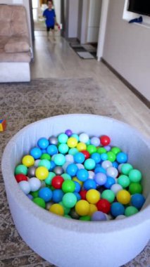Sevimli aktif çocuk odaya giriyor. Bebek, plastik toplara doldurulmuş kuru havuza atlıyor. Dikey video.