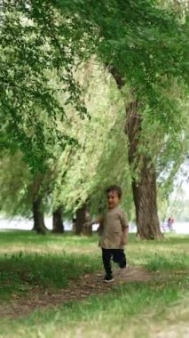 Mutlu gülümseyen küçük çocuk parkın önünden geçiyor. Aktif erkek bebek yazın yürüyüşe çıkıyor. Dikey video.