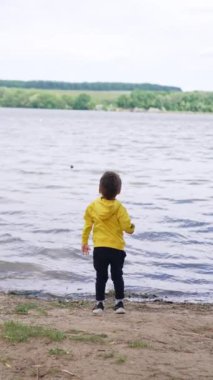 Küçük çocuk nehre doğru yürür ve bir taş atar. Mutlu çocuk arkasını dönüp el çırpıyor ve kameraya doğru koşuyor. Dikey video.