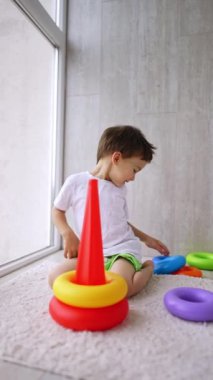 Yerde oturan sevimli, gülümseyen bebek piramit yapıyor. Sevimli çocuk evde, pencerenin yanında oynuyor. Dikey video.