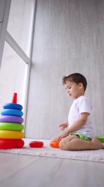 Pencerenin önünde oturan sevimli çocuk bir piramit inşa ediyor. Evde oynayan çocuğa düşük açılı bakış açısı. Dikey video.