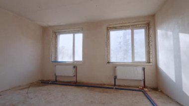 Altında iki pencere ve radyatör olan boş bir oda. Ampulle aydınlatılan gri duvarlarla karanlık koridora giriyoruz. Yeniden yapılanma kavramı.