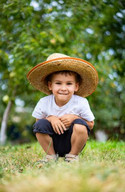 Young boy enjoying a sunny day in the garden. A cheerful boy wearing a straw hat squats in a grassy area surrounded by trees, enjoying a sunny outdoor day. clipart