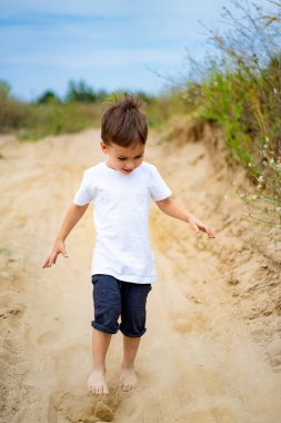 Genç çocuk dışarıda kumlu yolda yalınayak oynuyor. Beyaz gömlekli genç bir çocuk güneşli bir günde çıplak ayakla oynarken kumlu bir yolu keşfediyor..