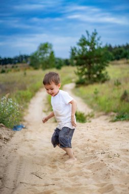 Güneşli bir günde kumlu yolda oynayan bir çocuk. Genç bir çocuk yeşilliklerle çevrili kumlu bir patikada neşeli bir şekilde koşuyor, güneşli bir öğleden sonranın tadını çıkarıyor..