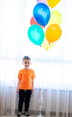 Evin içinde renkli balonlarla ayakta duran bir çocuk. Turuncu tişörtlü bir çocuk elinde beyaz perdeli bir odanın içinde bir demet balon tutarken gülümsüyor..