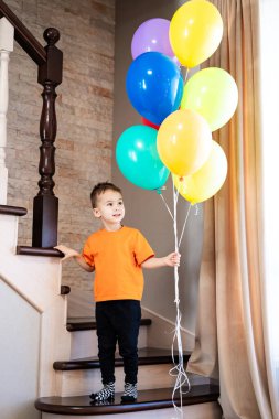 Çocuk neşeyle renkli balonlar tutuyor. Turuncu tişörtlü neşeli bir çocuk elinde renkli balonlarla merdivenlerin üzerinde duruyor..