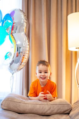 Çocuk sıcak oturma odasında balonlarla kutlama yapıyor. Turuncu tişörtlü gülümseyen bir çocuk, sıcak bir oturma odasında renkli balonlarla doğum gününü kutluyor..