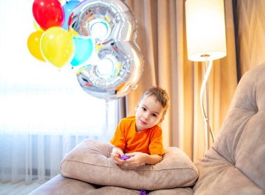 Çocuk 3. doğum gününü balonlarla kutluyor. Turuncu gömlekli bir çocuk üçüncü doğum gününü renkli balonlar ve kapalı alanda rahat yastıklarla kutluyor..