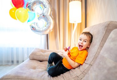 Çocuk doğum gününü balonlarla kutluyor. Neşeli bir çocuk kanepede oturmuş, etrafında canlı balonlar ve yüzünde kocaman bir gülümsemeyle doğum gününün tadını çıkarıyor..