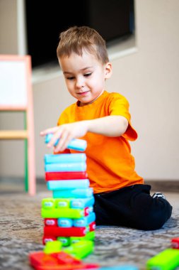 Child stacking colorful building blocks indoors. Young child in an orange shirt plays with colorful building blocks while sitting on the floor in a bright room. clipart