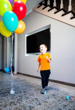 Evin içinde renkli balonları olan mutlu bir çocuk. Turuncu tişört giyen mutlu bir çocuk, sıcak bir mekanda canlı balonlar tutarken koşar..