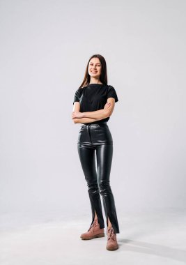 Genç bayan kendine güvenen bir şekilde şık kıyafetler içinde poz veriyor. Genç bir kadın kollarını kavuşturmuş modern siyah pantolonları ve stüdyo ortamında sade bir bluzu sergiliyor..