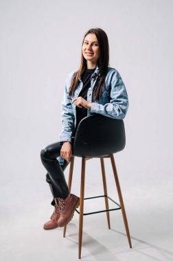 Stüdyo ortamında taburede poz veren genç bir kadın. Genç bir kadın, parlak bir stüdyoda kot ceket, siyah bluz ve şık çizmeler giyerek bir tabureye güvenle oturur..