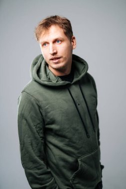Kapüşonlu genç adam, sade bir geçmişi var. Açık kahverengi saçlı ve kapüşonlu mavi gözlü bir adam stüdyoda düşünceli bir şekilde yan tarafa bakar..