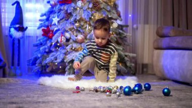 Güzel beyaz erkek bebek süslü Noel ağacının yanındaki tüylü paspasın üzerine uzanıyor. Çocuğun yanında renkli oyuncaklar var..