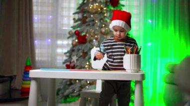 Noel Baba şapkalı sevimli beyaz erkek bebek masanın üstünde duruyor. Çocuk bardaktan bir kalem seçiyor ve çizmek için oturuyor. Bulanık arkaplanda Noel ağacı.